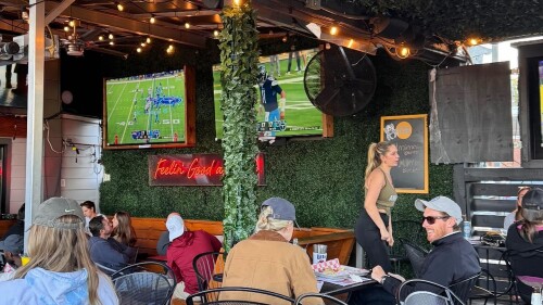 A group of people sit on a patio of a covered restaurant watching a football game on a monitor.