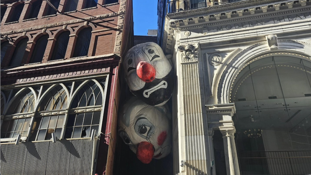 Two large, inflatable clown heads sandwiched in an alley between the ornate architecture of two buildings.