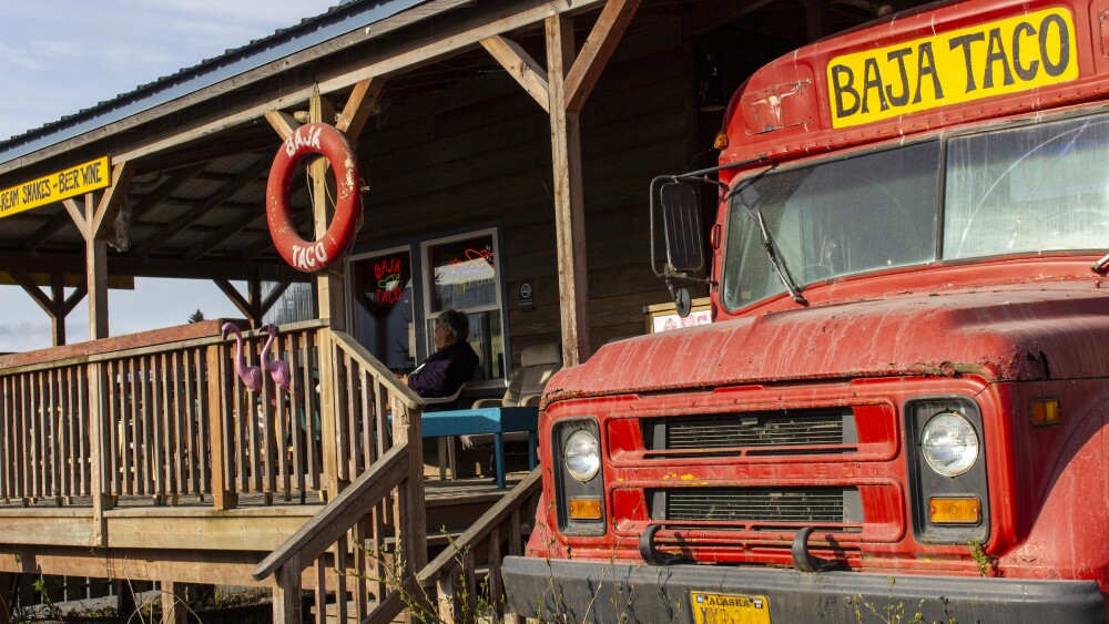 A red school bus with an Alaska license plate holds a sign saying Baja Taco, with an attached wooden patio and building.