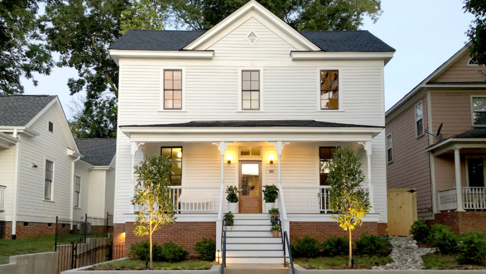 Charming white two-story house with a front porch, warm lights, and potted plants. Evening setting with trees and neighboring houses in view.
