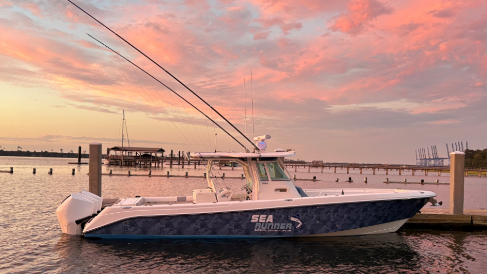 A sleek fishing boat named "Sea Runner" is docked at a marina during a stunning sunset. The sky is painted in vibrant hues of pink, orange, and purple. Calm waters reflect the colors, and long fishing rods extend skyward. The scene exudes serenity and maritime adventure.