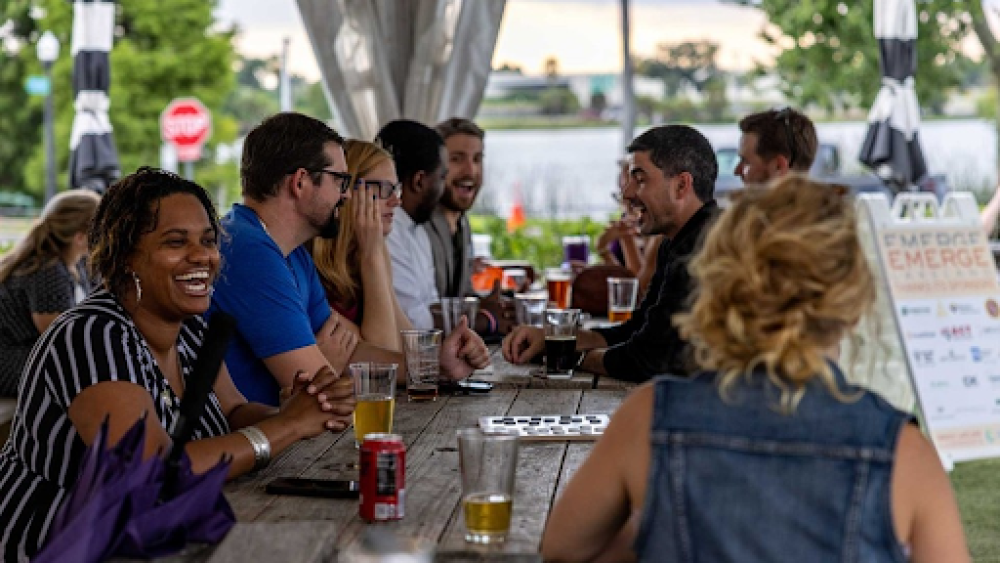 A group of young professionals laugh, talk, and drink beer at a long table outside. A sign reading "emerge" is visible in the background.