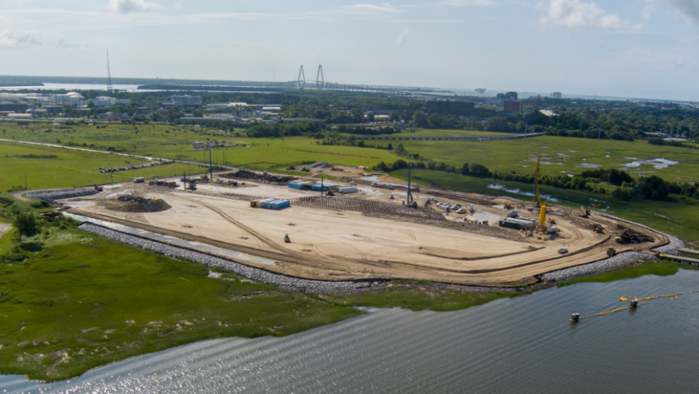The photo above shows the Magnolia Landing Construction site. There's a large brown dirt plot with excavators and equipment, surrounded by lush greenery and a waterfront — you can see the Ravenel Bridge in the background.