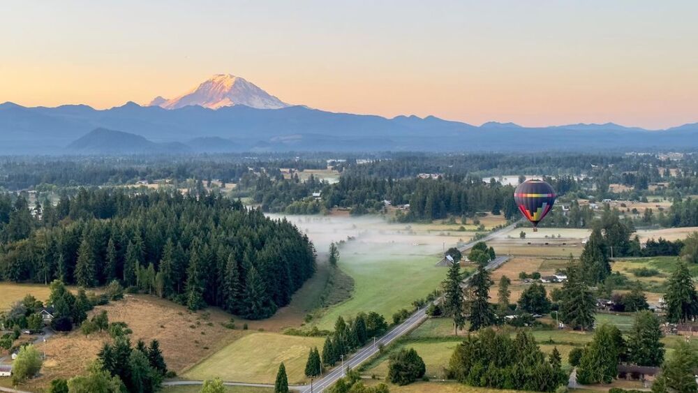 A hot air balloon with a rainbow geometric pattern hovers above a rural residential area while Mt. Rainier stands proud in the distance during a sunrise trip.