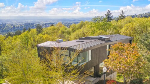 An aerial photos shows a modern house on the edge of a grove of deciduous trees with a valley stretching away below into the distance.