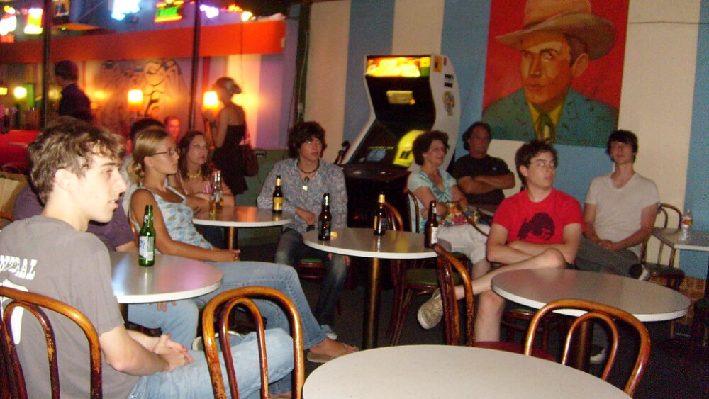 A group of people sit at small round tables in a dimly lit bar, watching something off camera. Most of them hold beer bottles and appear relaxed and focused. The bar features colorful neon lights, an arcade machine, and a large, vibrant painting of a man in a cowboy hat on the wall. The atmosphere is casual and retro.