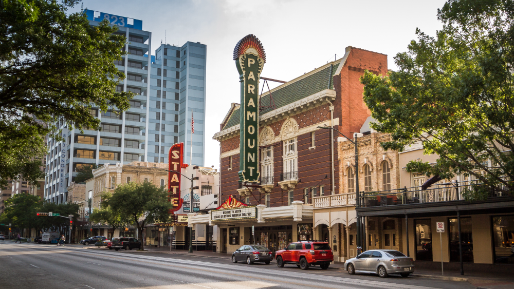 The exterior of the Paramount Theatre in Austin, Texas; the marquee hangs on the entrance of the historic brick building, along with a vertical dark green sign that says "Paramount". A few cars line the street in front of the venue.