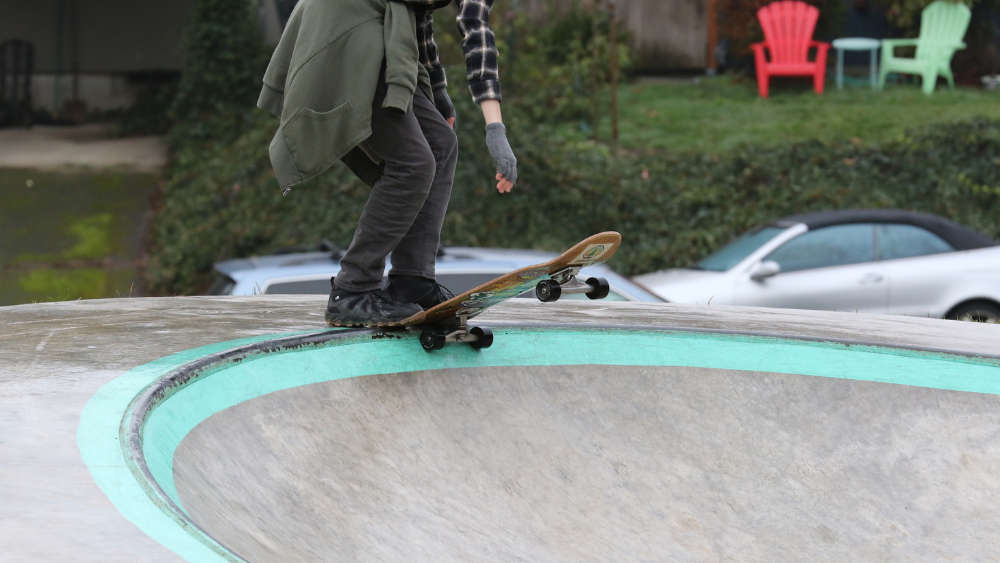 A skater waits with their skateboard resting on and out from the lip of a concrete bowl at the skate area in Error Heights Park.