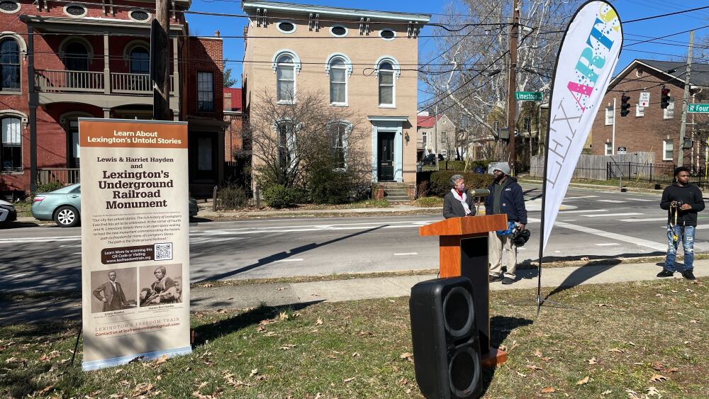 An outdoor space with a podium, speaker, a sign that reads, "Learn About Lexington's Untold Stories" and a flag that says, "LexArts."