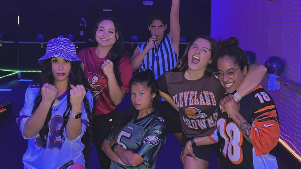 Instructors at Rock It Cycle and Fitness are dressed in football outfits. There is purple lighting and the five instructors are holding fun poses, including clenched fists, with one referee blowing a whistle.