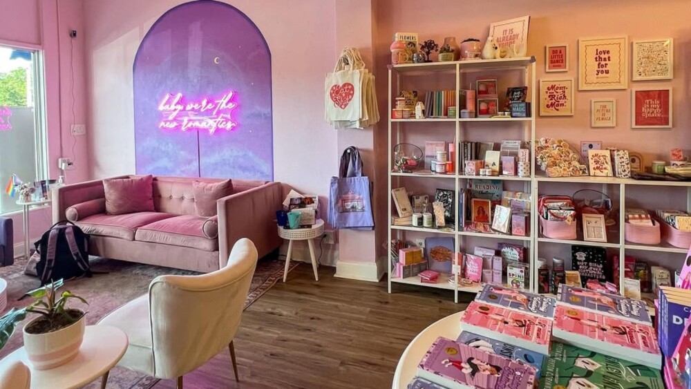 Cozy pink-themed interior of The New Romantics bookstore with a velvet sofa, small tables, and shelves filled with books, candles, and gifts. A neon sign on a purple wall reads, “baby we’re the new romantics.” The space has a playful and inviting atmosphere.