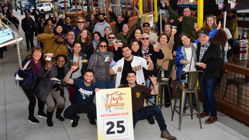 A group of people gather with drinks and food in hand, smiling and toasting to Taste of Third in Chula Vista.