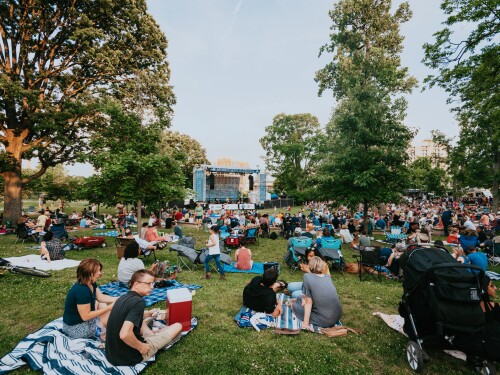 A crowd of people lounge on blankets on a grassy lawn in front of a stage at a live music event.