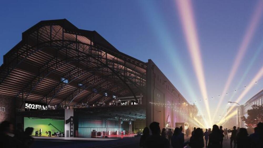 A rendering of a large, open-ended building with multiple film sound stages inside, along with an open boulevarde running along its right-hand wall, which is filled with people + large, multicolored spotlight-style lights shining into the sky.