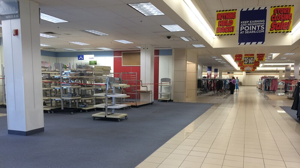 A store interior with multiple brightly-colored signs hung from the ceiling that say things like "Store closing sale," "Nothing held back," and "25%-60% off." The main portion of the store pictured has various empty shelves on wheels, with a few people seen shopping clothing racks in the background.