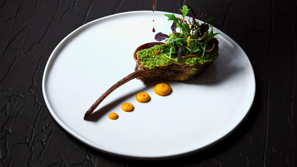 Sauce is poured over a decorated lamb chop on a white plate on a dinner table.