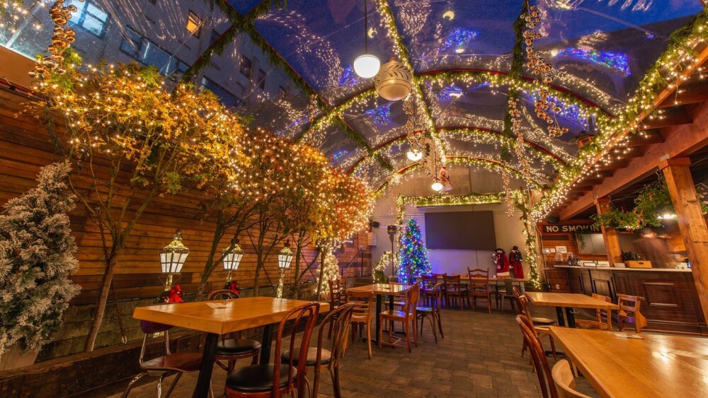 A cafe with a convex glass roof is decorated with warm string lights, lanterns, and holiday greenery. Inside are tables and mismatched chairs. 