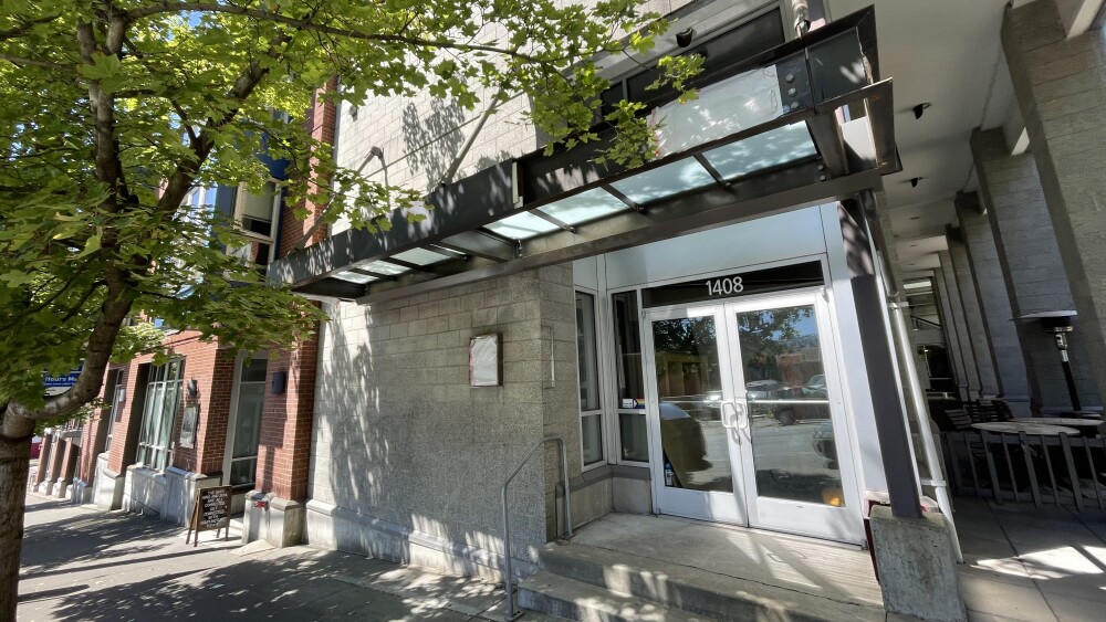 The corner of a building features silver metal doors and concrete steps, framed by the leafy branches of a tree. The storefront doesn't bear any signage or markings, but it will soon be the home of Stitch Cafe.