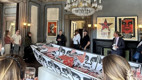 This photo depicts a gathering of people inside an ornate, historical room with vintage decor. The room features distressed walls with decorative molding, a grand chandelier with multiple candle-style lights, and a black fireplace adorned with framed artwork. The artwork includes iconic "Obey" prints associated with Shepard Fairey, featuring bold graphic designs in red, black, and white.
In the center of the room, a striking art installation is displayed — a boat covered in Fairey's signature designs, including the word "OBEY" and stylized motifs. Several people are present, some are engaged in conversation, while others are observing the artwork.