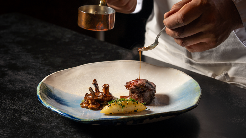 A person pours a brown, gravy-like sauce on top of a small piece of beef plated at an upscale tasting-menu-only restaurant in San Diego