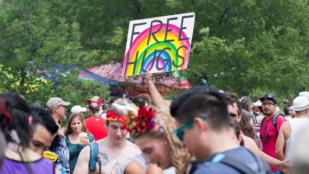 A crowd of people wearing colorful clothing with a rainbow sign reading "Free Hugs" in the air.