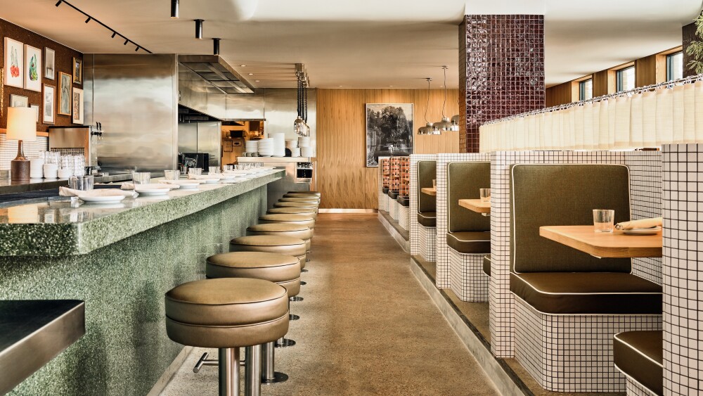 A modern restaurant interior featuring a long green terrazzo bar with round leather stools, open kitchen in the background, and a row of two-person booths with olive green cushions and white tiled walls. Warm lighting, floor-to-ceiling windows, and wood accents create an airy, inviting atmosphere.