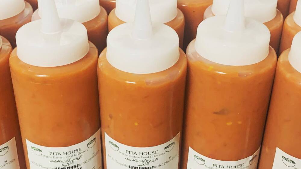 Several rows of clear bottles that contain an orange sauce. On the front of the bottles is a white sticker that says "Pita House" and "Homemade Hot Sauce."