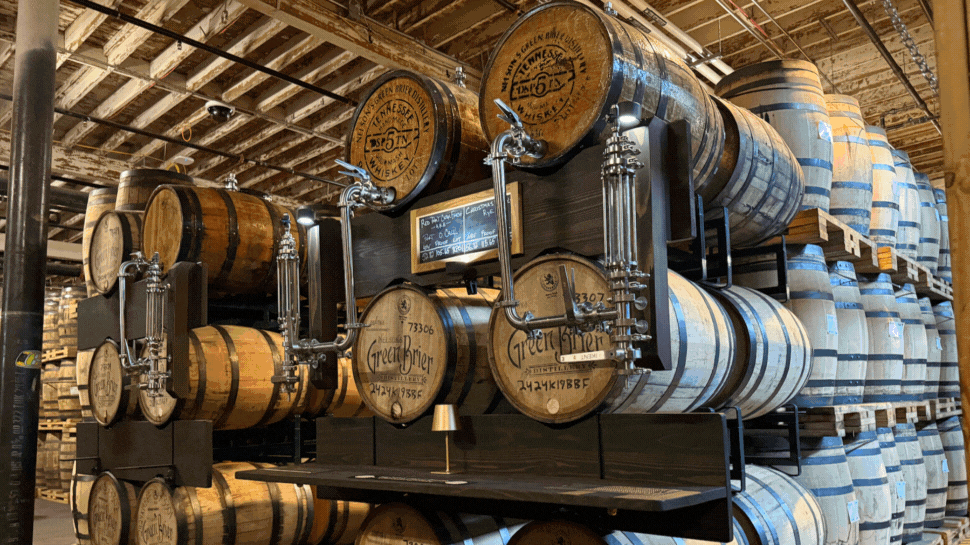 Photo 1: Wooden barrels stacked in a spacious distillery warehouse. Metal taps attached to some barrels. Photo 2: A hand holds a whiskey glass in focus, showing amber liquid inside. Blurred background of people at a tasting, creating a cozy, intimate atmosphere. Photo 3:  A dining table with a rustic wood finish features a cocktail, soft pretzels with cheese dip, condiments, and a plate of fries with a sandwich. Photo 4: Sidewalk view of a gray brick distillery with a sign for "Distillery Tastings & Gift Shop." The sky is clear blue, creating an inviting atmosphere. Photo 5: Large stainless steel brewing tanks are lined up in a modern brewery, with informational signs in front. A person in a beige jacket observes the process.