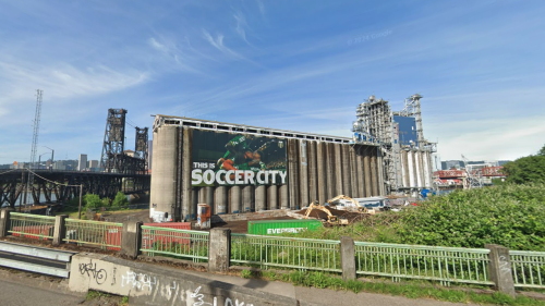 A Google Street View image taken in 2024 of a large industrial building with a billboard of a soccer player that reads "This is Soccer City." The Steel Bridge is visible in the background.