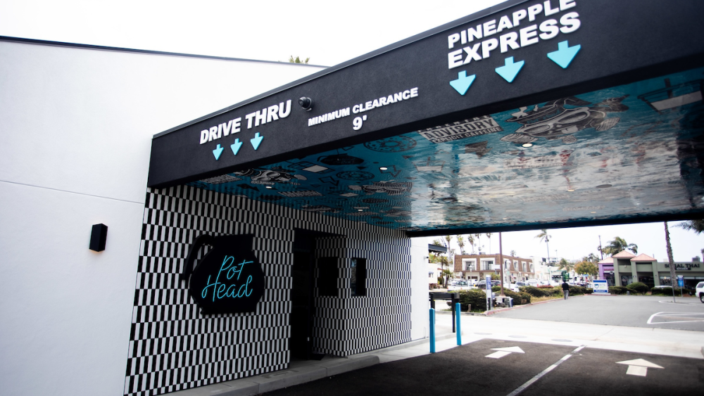 A drive-thru Spill the Beans coffee shop in Ocean Beach; the awning is filled with blue and black illustrations and the drive-thru wall features a playful sign that reads "Pot Head."
