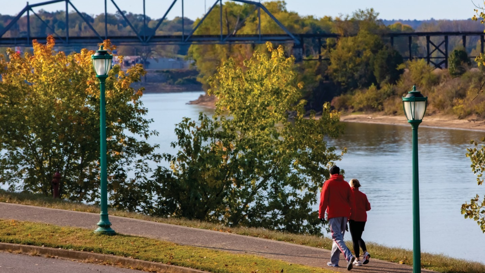 Two people in warm athletic clothing walk a paved path along the river towards a bridge surrounded by lush greenery turning colors to fall.