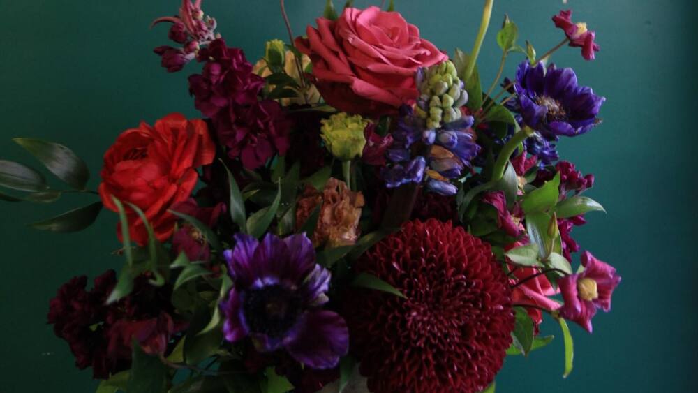 This flower arrangement from Verde & Co. has leaned into a dramatic color palette with red roses, burgundy mums, and a deep emerald background.