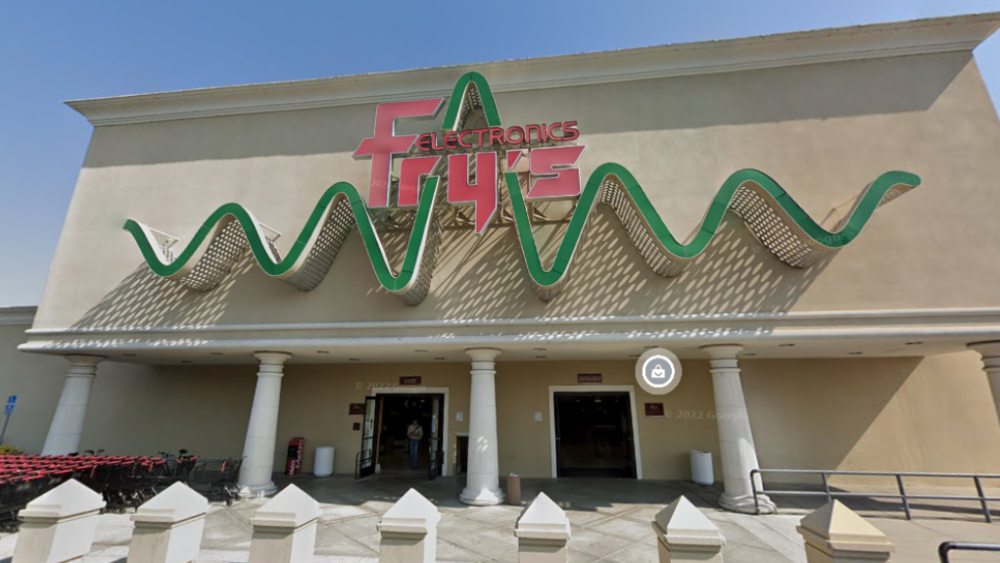 The front of a big beige Fry's Electronics building. The store's logo is red, sitting in the middle of a dark green waveform. The doors are open, and it appears lively.