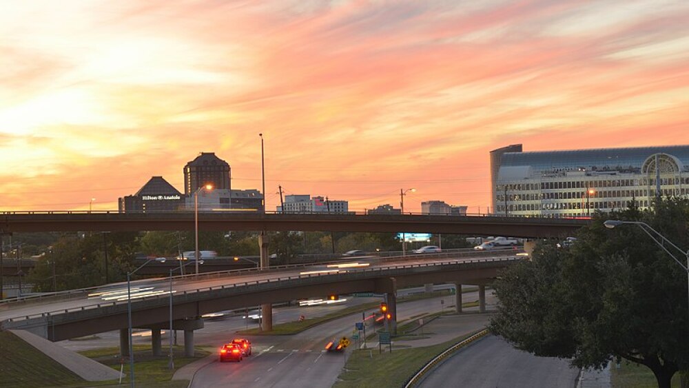 A sunset with hues of orange, pink, purple and blue set behind two overpasses. The tops of several tall buildings are visible behind the roads.