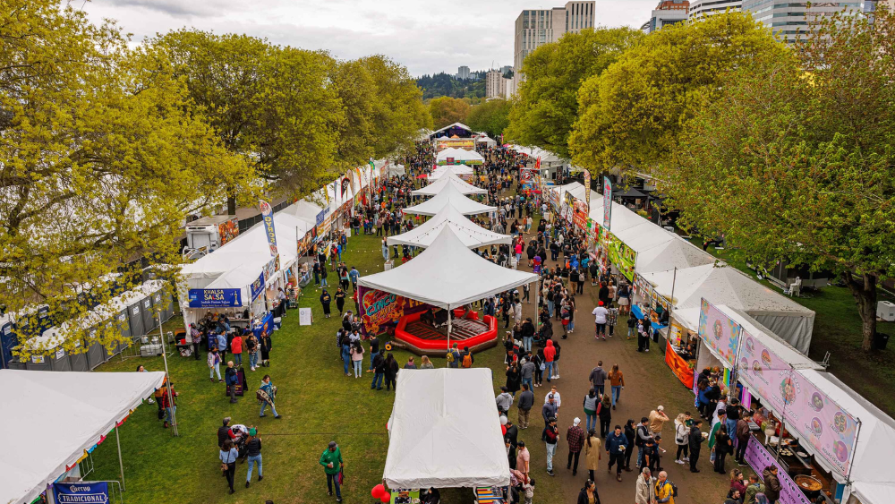 Looking down on three rows of white festival tents stretching into the distance, surrounded by trees with the Portland skyline as a backdrop. People mill about the tents.