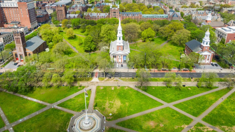 An aerial view of the city of New Haven, Connecticut. We see a large brick clock tower, green fields, and other city buildings surrounding it.