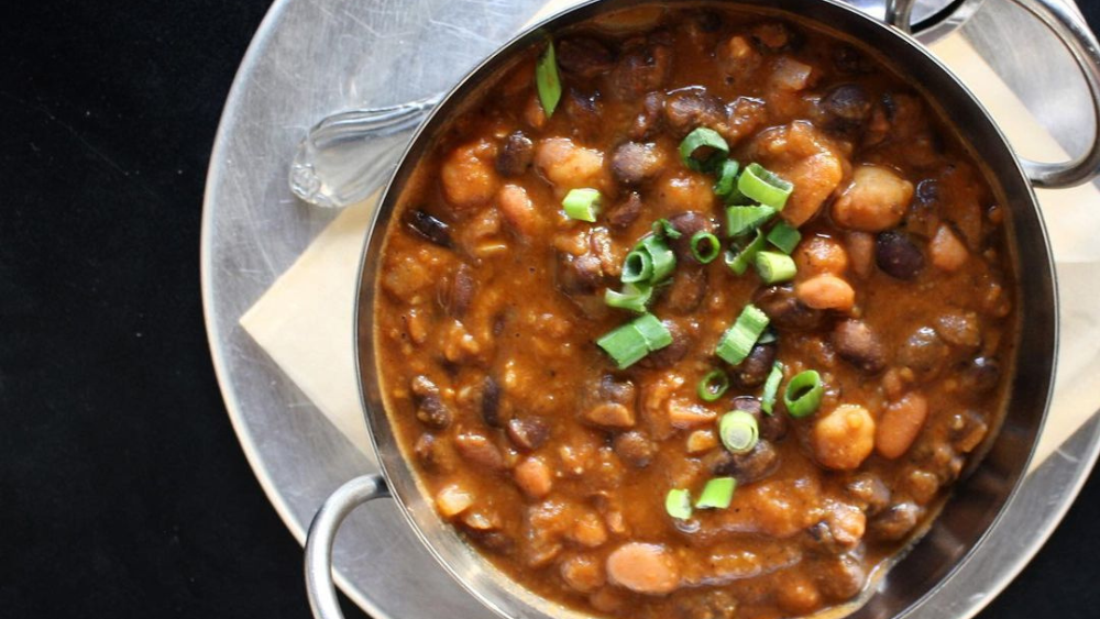 A bowl of three bean stew sits on a black table top, pictured from above.