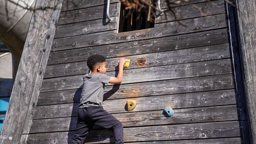 A child climbs rockwall fixtures up a wooden wall, while his mother braces his feet.