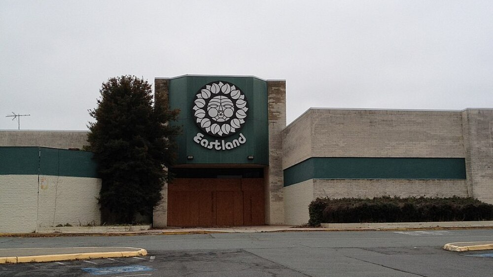 The exterior of a building behind an empty parking lot on a cloudy, grey day. The building is made of dirty white and green brick, with a sign that says "Eastland" on the outside.