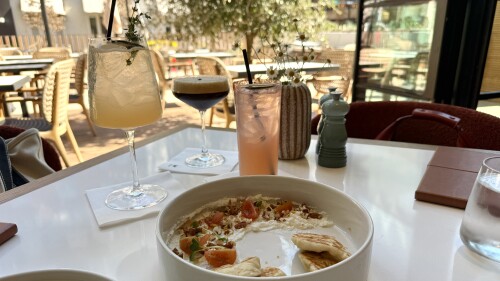 A table set at an outdoor restaurant with a bowl of whipped cheese topped with diced tomatoes, herbs, and nuts, accompanied by pieces of toasted flatbread. Three cocktails are in the background: a pale yellow drink in a wine glass with a sprig of thyme, a dark espresso martini in a coupe glass, and a pink beverage in a tall glass with a black straw. The setting features wicker chairs, a potted plant, and natural light streaming through open windows.
