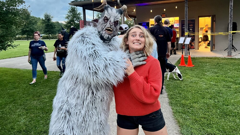 A person dressed as the Pope Lick Monster, wearing a furry costume and goat horns, pretends to choke City Editor Katie. The photo has a playful and staged air about it.