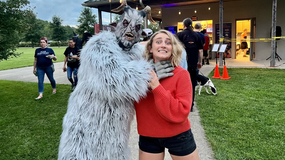 A person dressed as the Pope Lick Monster, wearing a furry costume and goat horns, pretends to choke City Editor Katie. The photo has a playful and staged air about it. 