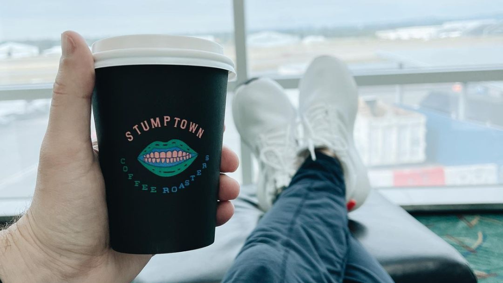 A hand holds a cup from Stumptown Coffee Roasters in the Portland International Airport. Crossed legs and the runway are in the background.