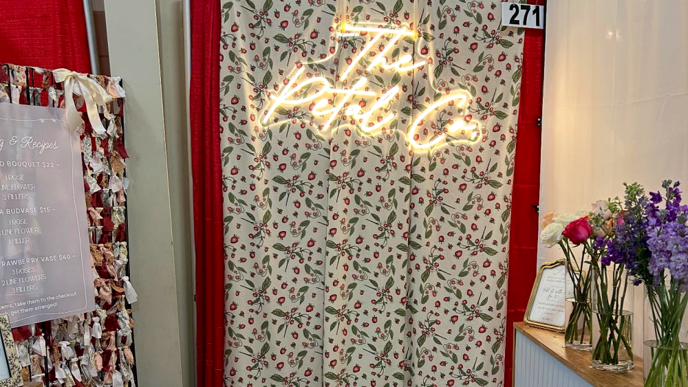 A small indoor booth at a festival with a flower bar and a neon sign reading "The Petal Co."