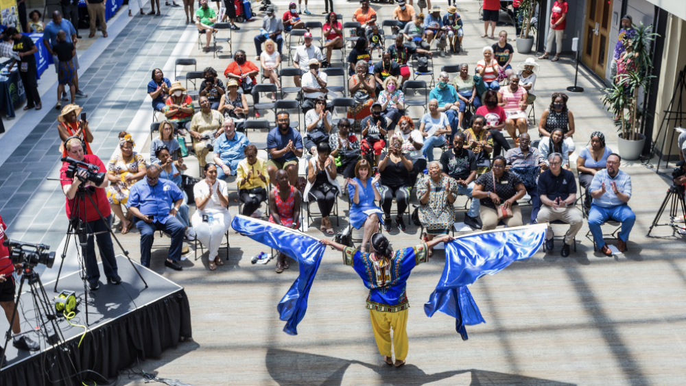 A group of people sitting in chairs watches a person holding blue flags outstretched in either arms. The presentation is being filmed by staff on the left side on a stage.