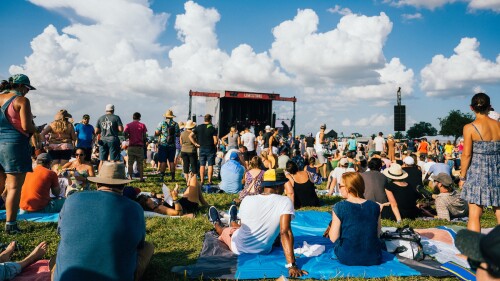 People laying and standing on a lawn in front of a stage.