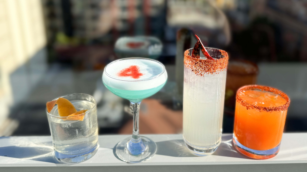 Four cocktails sit on a ledge — one is clear with a fruit twist, one is turquoise blue with the number 4 in the froth, one is light yellow with an orange rim, and one is bright orange.