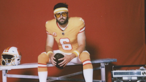 A vintage-looking photo of Bucs quarterback Baker Mayfield wearing an orange creamsicle football uniform. His retro glasses have orange lenses, and he's wearing an old-school headband. There is a boombox next to the bench he's sat on, he's looking at the camera with a serious expression, and the background is a plain red sheet.