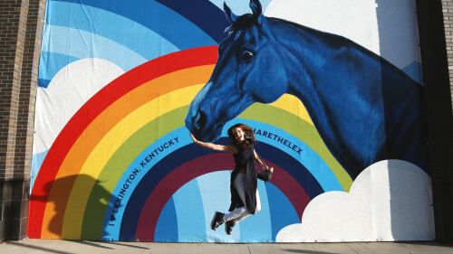 A woman jumps in front of a tapestry featuring a rainbow and horse, with text that reads "Lexington, Kentucky #sharethelex"