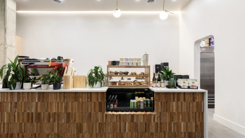 A bright coffee counter. There are wooden features, drinks in a fridge, baked goods in a glass case, and no one in the shot.